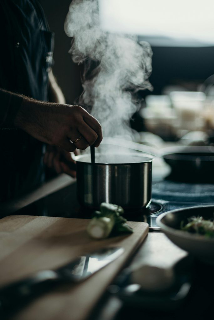 A person stirring a steaming pot on an electric stove with kitchenware spread out.